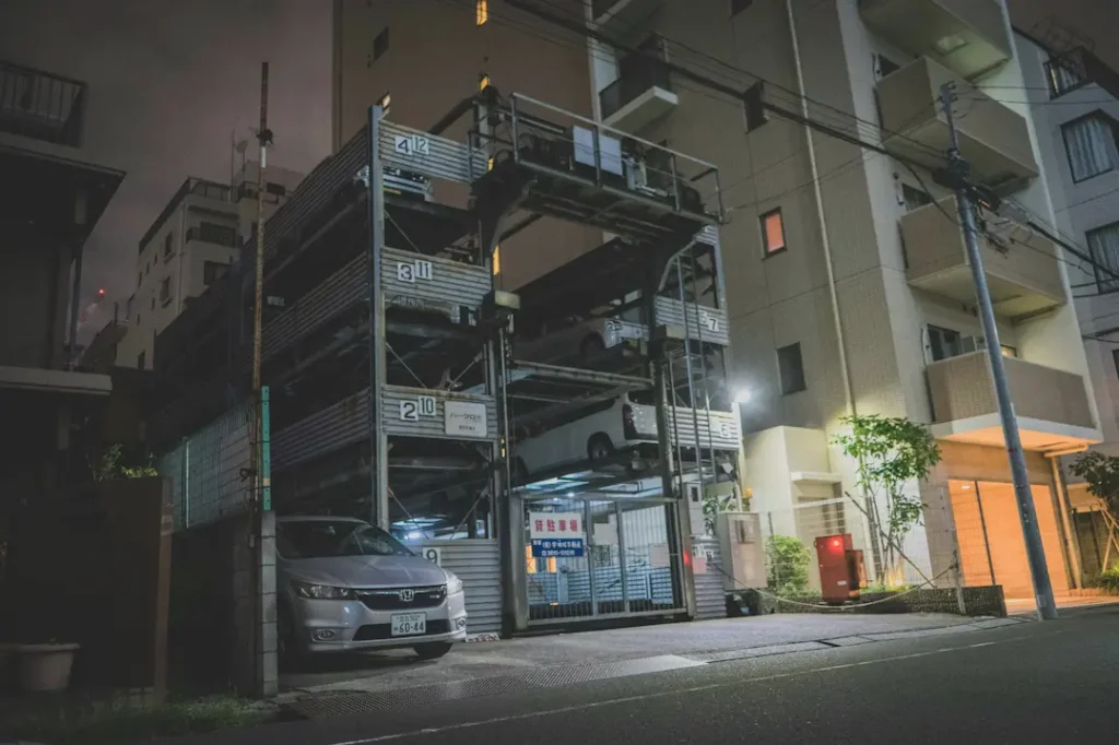 Nocna ulica w japońskim mieście z wielopoziomowym parkingiem mechanicznym, zaparkowanymi pojazdami, budynkami mieszkalnymi, liniami energetycznymi i łagodnie oświetlonymi wejściami.